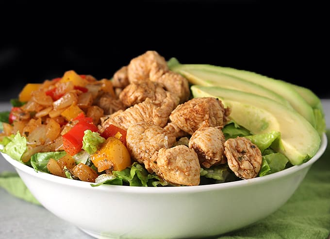 A bowl of lettuce, chicken, peppers, and avocado with a black background. 