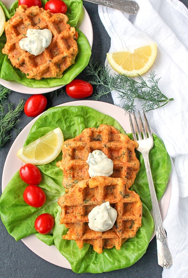 A plate with two salmon cake waffles on a lettuce bed with a lemon slice and tomatoes on the side. 