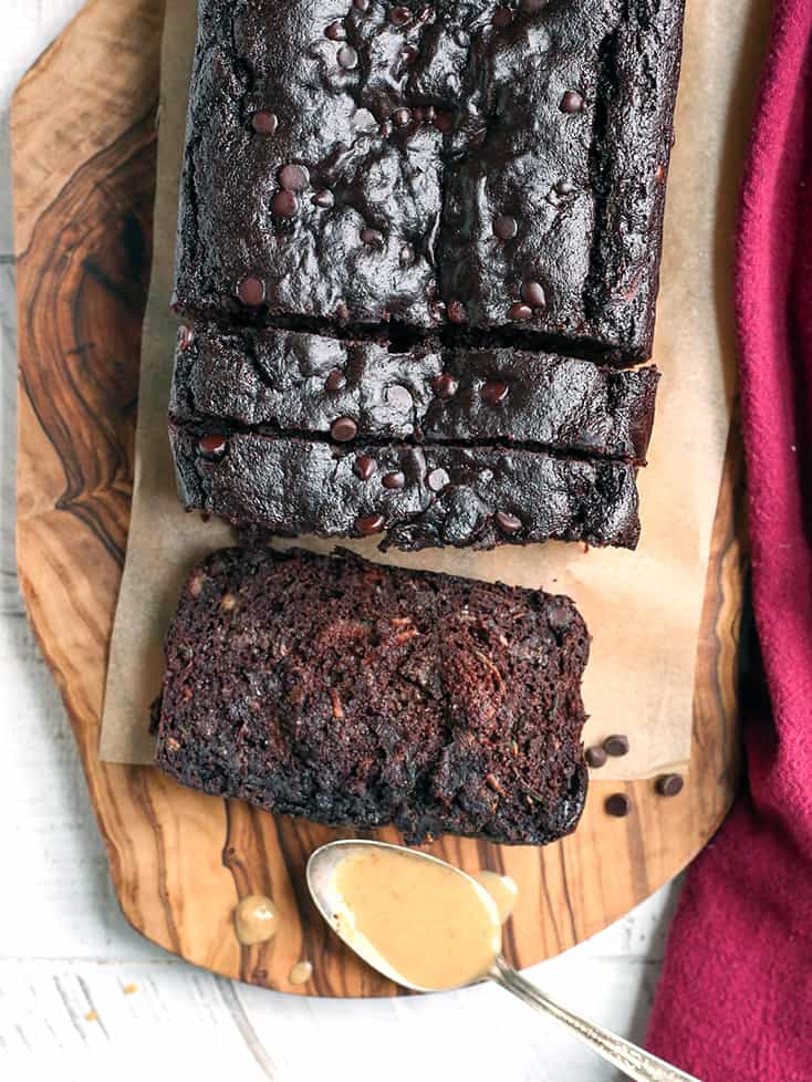 a loaf of chocolate paleo vegan zucchini bread on a wooden cutting board. Sliced with a piece laying flat