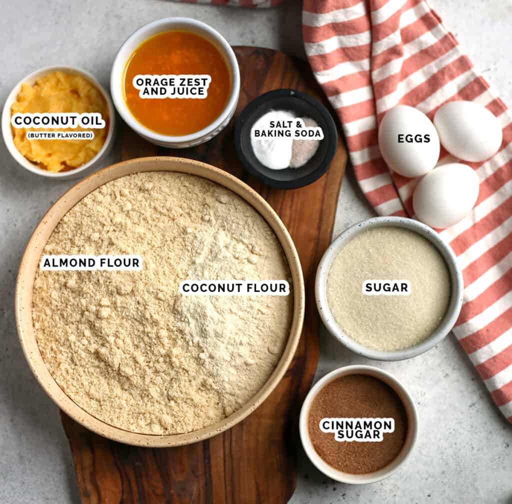 an ingredient image showing coconut oil, orange juice and zest, salt, baking soda, eggs, sugar, almond flour, coconut flour, and cinnamon sugar all in separate bowls. 