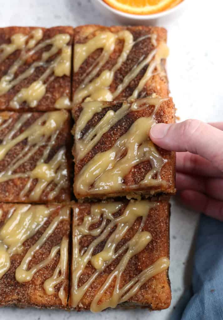 a hand reaching for a piece of paleo cinnamon coffee cake, showing the top  of the cake with a glaze. 