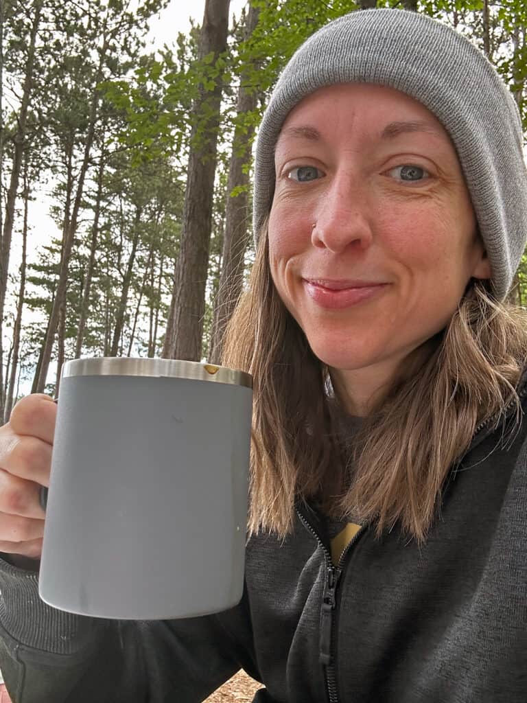 a white woman with a knit hat on holding a cup of coffee outdoors. She is smiling and the trees are behind her. 
