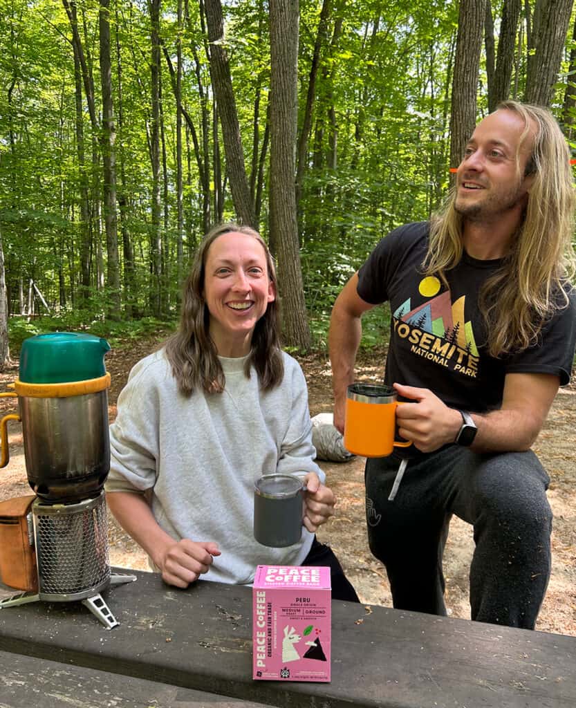 a man and woman enjoying coffee while camping sitting at a picnic table. Woods in the background. 