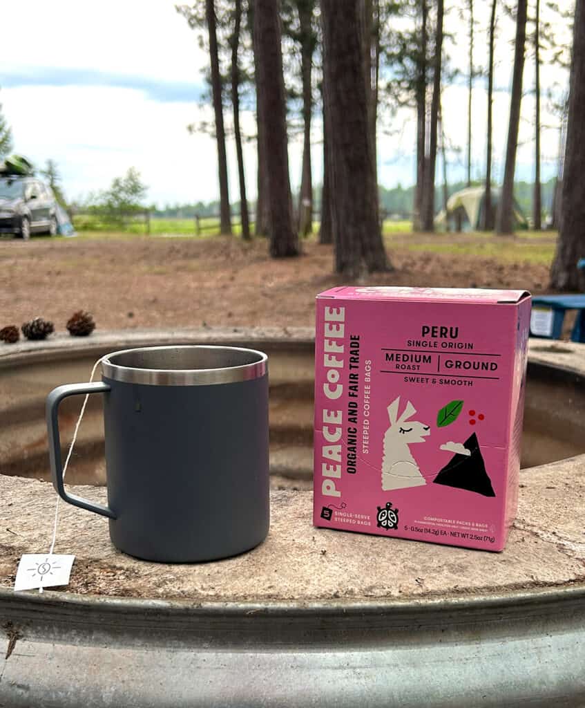 a coffee mug sitting on the edge of a fire pit with a box of steeped coffee next to it. A tent and river in the background. 