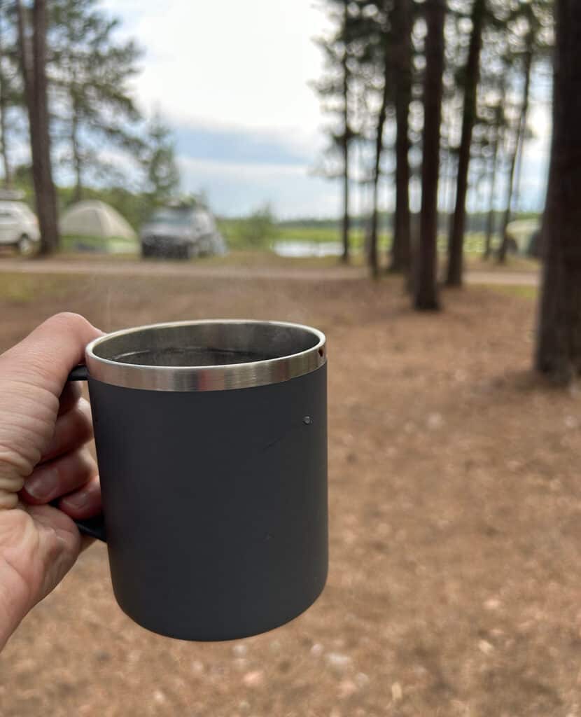a hand holding a camping cup of coffee with tents, trees and a river in the background. 