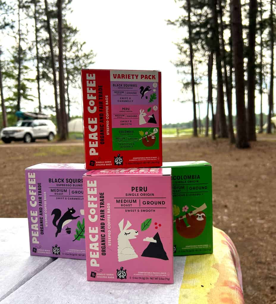 Four boxes of steeped coffee sitting on a picnic table. Trees, a tent and a river in the background. 