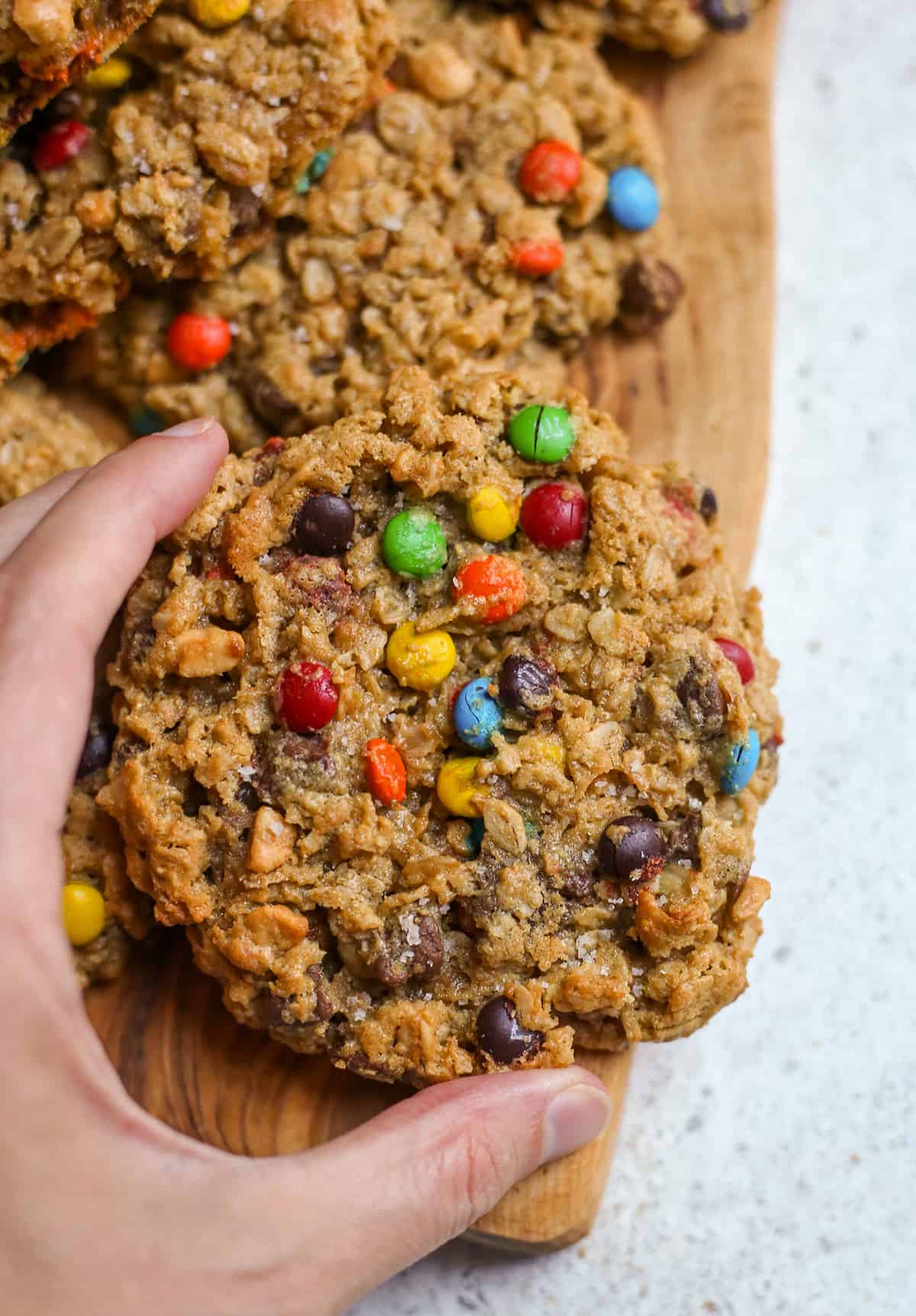 hand grabbing a monster cookie that is on a wooden cutting board. 