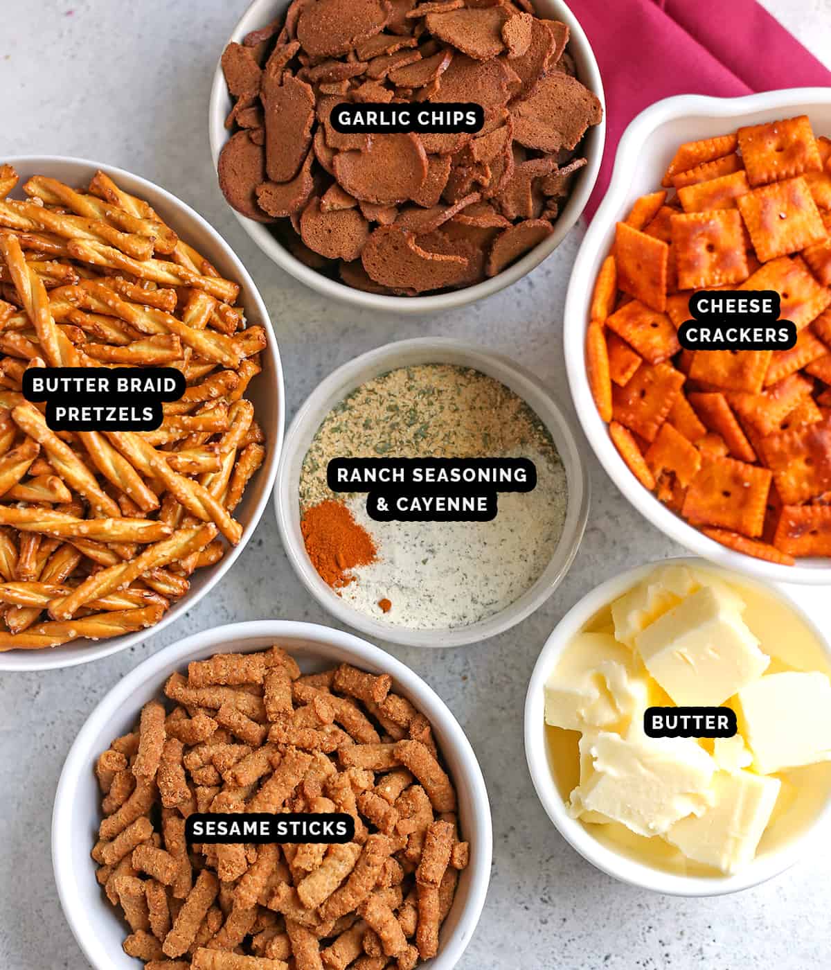 Ingredients for snack mix all in their own separate bowls and labeled- pretzels, garlic rye chips, cheese crackers, sesame sticks and seasoning. 