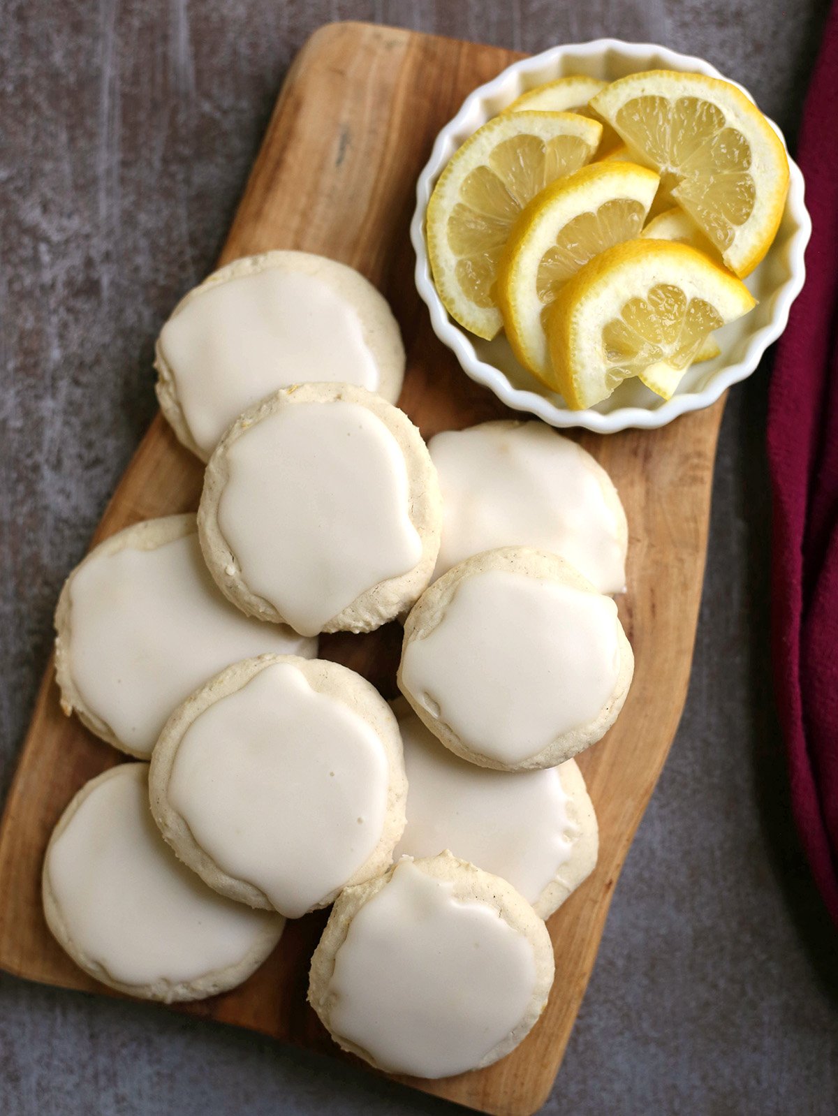 A stack of lemon cookies with frosting on a wooden cutting board and a little dish with sliced lemons in the top right corner.